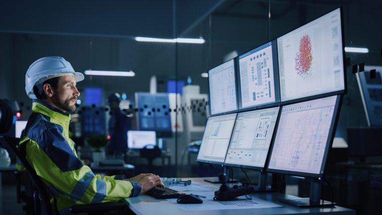 Modern Factory: Facility Operator Controls Workshop Production Line, Uses Computer with Screens Showing Complex UI of Machine Operation Processes, Controllers, Machinery Blueprints